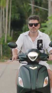 Dikey video. Beyaz Gömlekli Adam Viraj Yolu 'nda yavaşça scooter kullanıyor. Palmiye ağaçlarıyla aynı hizada. Önden hafif açılı, Yavaş Hareketli, Tropikal Sürücü Konsepti.