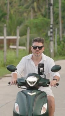 Dikey video. Güneş Gözlüklü Adam 'ın Yakın Çekimi ve Beyaz Gömlekli Scooter Tropikal Yol Boyunca, Orman Arkaplanı, Yavaş Hareketli Ön Çekim, Ada Özgürlüğü Konsepti