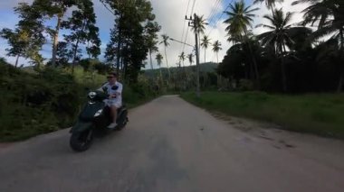 Scooter 'daki adam Palm Trees' in altından geçiyor. Karışık Toprak ve Asfalt Yolundaki Kameraya doğru gidiyor. Drone Side Açı Vuruşu, Tropikal Özgürlük Konsepti.