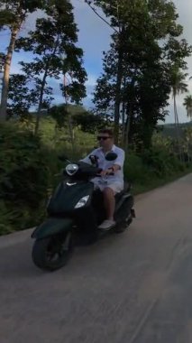 Dikey video. Scooter 'daki adam Palm Trees' in altından geçiyor. Karışık Toprak ve Asfalt Yolundaki Kameraya doğru gidiyor. Drone Side Açı Vuruşu, Tropikal Özgürlük Konsepti.