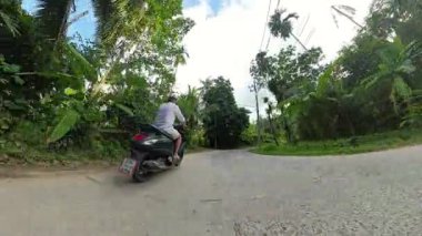 Palm Road boyunca Scooter süren adam, düşük güneş ışığı, geniş açılı arka lastik seviyesi, günbatımı tropikal yolculuk konsepti.