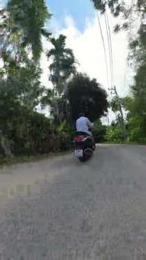 Dikey video. Palm Road boyunca Scooter süren adam, düşük güneş ışığı, geniş açılı arka lastik seviyesi, günbatımı tropikal yolculuk konsepti.