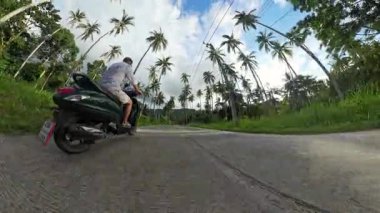 Sunset Light 'ta Lush Palm Grove' da Scooter süren adam, Sol Taraftan Geniş Mercek İzleme Çekimi, Tropikal Sürüş Konsepti