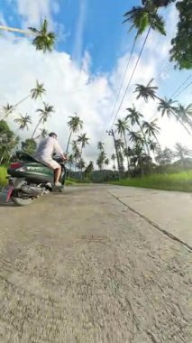 Dikey video. Sunset Light 'ta Lush Palm Grove' da Scooter süren adam, Sol Taraftan Geniş Mercek İzleme Çekimi, Tropikal Sürüş Konsepti