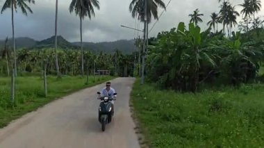 Palm Trees ve Bulutlu Dağlar Arkaplanda Dönen Kırsal Yol boyunca Scooter süren Adam, Orta Yol, Tropikal Seyahat Yolu