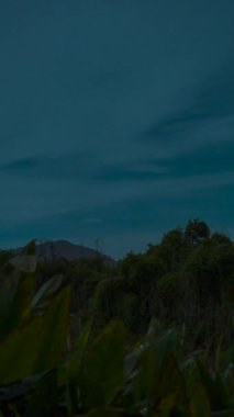 Dikey video. Yoğun Yeşillik ve Siluetli Dağ Sırtı ile Tropikal Zaman Hızı Sahnesinde Gece Dönüşümleri Gibi Fırtınalı Bulutlar Yavaşça Dönüşüyor