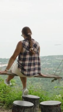 Dikey video. Cliffside Bakış Açısı 'ndan Tahta Salıncak Üzerinde Oturan Kadın Ağaçlar, Orta Salıncak, Doğa Konsepti