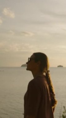 Dikey video. Güneş Gözlüklü Kadın Gün Batımında Sahilde Yürüyor Su Üzerine Altın Yansımalar, Huzurlu Gezinti Konseptiyle