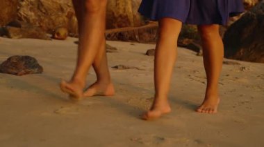 Gün batımında birlikte bir Rocky Plajı boyunca çıplak ayakla yürüyen çift. Bacakları ve günlük kıyafetleri üzerindeki ılık ışıkla romantik sahil yürüyüşü kavramı.