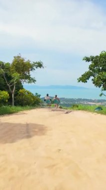 Dikey video. FPV İnsansız Hava Aracı Hızlı Uçuyor Gün Işığında Yükselen Ağaçlarla çevrili Yükseltilmiş Ocean ve Coastal Hills 'in Manzarasına Bakan Benç Manzarasında Oturan Bir Çift Geçiyor