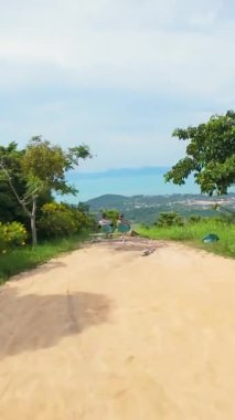 Dikey video. FPV İHA, Açık Mavi Gök Altındaki Mesafede Tepeler ve Sahil Kenti ve Okyanus Manzaralı Yoğun Yeşil Ağaçlar Üzerine Hızla Aşağı Dalıyor