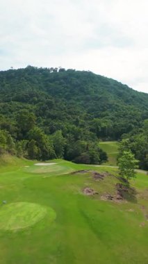 Dikey video. FPV İHA, Parlak Gök Altında Yoğun Tropikal Orman ve Tepeler Arasında Lush Golf Sahasında Uçuyor