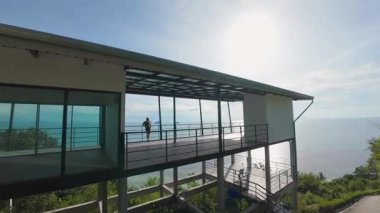 FPV insansız hava aracı geçitten geçip hızla ağaçlara ve aşağıdaki suya doğru inerken Okyanusa bakan Tepedeki Terasta Duran Adam