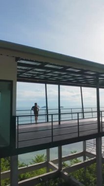 Dikey video. FPV insansız hava aracı geçitten geçip hızla ağaçlara ve aşağıdaki suya doğru inerken Okyanusa bakan Tepedeki Terasta Duran Adam