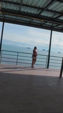 Dikey video. Hillside Yapısı Açık Terasında Duran Kadın Okyanusa Bakan FPV İHA Alttaki Ağaçlara Uçuyor