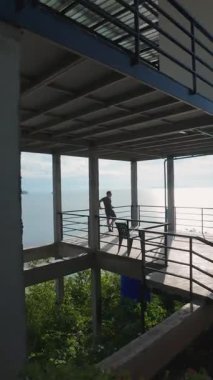 Dikey video. Beton Terasta Duran Adam Parlak Günışığında Okyanus Ufkusu 'na bakarken İnsansız Hava Aracı Düşük Açı' dan Uçuyor