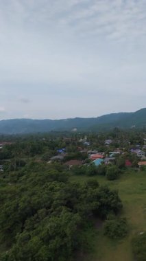 Dikey video. Yoğun Tropikal Ağaçlar ve Mavi Gök Altında Yuvarlanan Dağılmış Bulutlar ve Kırsal Yerleşim Konseptiyle çevrili Küçük Şehir Hava Manzarası
