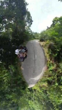 Dikey video. Dik Dar Yol 'da çift motosiklet sürüşü Etrafı Yoğun Ormanlarla çevrili Küçük Gezegen Efekti Genel Bakış Alanıyla çevrili