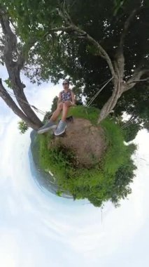 Dikey video. Ağacın Zevkini Çıkartan Kadın, Küçük Gezegen Kamera Efektiyle Lush Hilltop 'un Kıyısında Sallanıyor