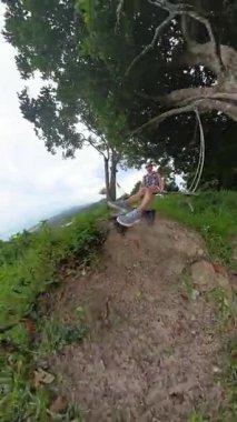 Dikey video. Ağaçta oturan kadın, okyanusa ve kıyılara tepeden bakan yüksek dağlarda sallanıyor.