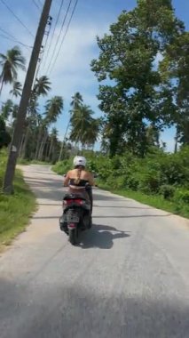 Dikey video. Dar yolda scooter süren Dar Palmiye Ağaçları Altında Parlak Mavi Gökyüzü, Yan Açı, Tayland Samui Adası Seyahat Konsepti