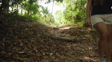 Tayland Ormanı 'nda Güneşli Tropikal Patikada Yürüyen Kadın Yaprak Kaplamalı Yerde Şapka ve Şort Giyiyor, Göz Seviyesi Çekimi, Doğa Yürüyüşü Kavramı