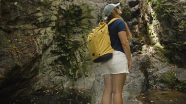 Sarı Sırt çantalı Kadın Tayland Ormanı 'nda bir uçurumun dibinde duruyor ve Kaya Surat' a Bakıyor, Yan Görünüm, Kamera Yavaşça İçinden Geçiyor, Engellerle Karşı Karşıya Gelme Kavramı