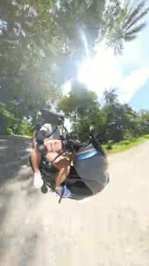 Dikey video. Erkek ve kadın, tropikal ormandan geçen beton bir yolda birlikte scooter sürüyorlar. Yandan çekilen 360 derecelik kamera, güneşli bir gün, Macera Yolculuğu Konsepti.