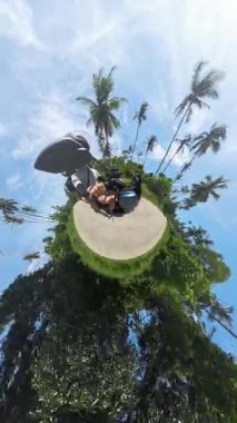 Dikey video. Palmiye ağaçları ve tropikal manzarayla çevrili küçük gezegen etkisindeki scooter kullanan çift, 360 derecelik kamera görüntüsü, parlak güneş ışığı, egzotik keşif kavramı.
