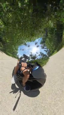 Dikey video. Küçük Gezegen etkisindeki tropikal arazide scooter kullanan çift, palmiyeler ve yeşillikler, güneşli hava, 360 derecelik manzara, açık hava gezisi kavramı...