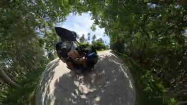 Erkek ve kadın, tropikal bir yolda, ağaçlar ve gölgelerle birlikte motosiklete biniyor, yan açılardan, güneşli havadan, doğa keşfi kavramından 360 derecelik kamerayla çekiliyor.