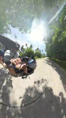 Dikey video. Erkek ve kadın, tropikal bir yolda, ağaçlar ve gölgelerle birlikte motosiklete biniyor, yan açılardan, güneşli havadan, doğa keşfi kavramından 360 derecelik kamerayla çekiliyor.