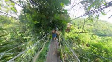 Yoğun yeşil orman üzerinde asılı bir köprüden geçen kadın geniş 360 derecelik bir tepe manzarası, canlı bitki örtüsü ve açık gökyüzü, Yağmur Ormanları Macerası Konsepti