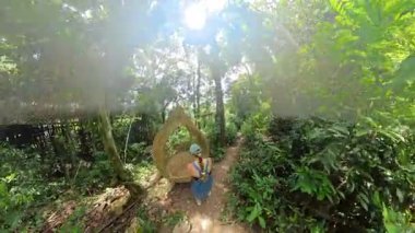 Kadın, tropikal orman ve ağaçlarla çevrili, dokuma şeklindeki gözyaşı damlası şeklindeki yuva koltuğuna tırmanıyor. 360 derecelik doğa görüntüsü, doğa etkileşimi konsepti...