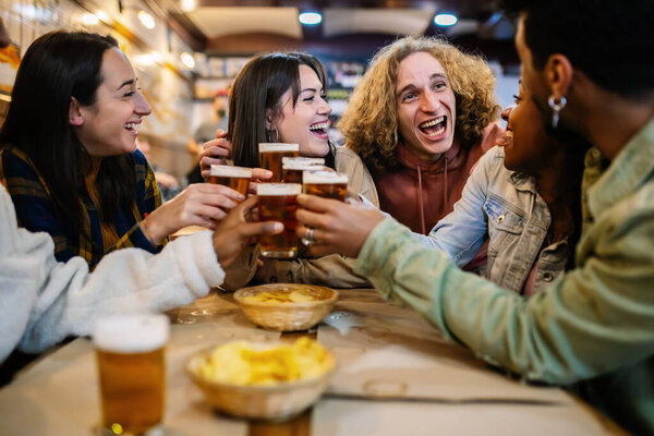 Young group of diverse best friends sitting in bar toasting with beer. Millennial happy student people celebrating together