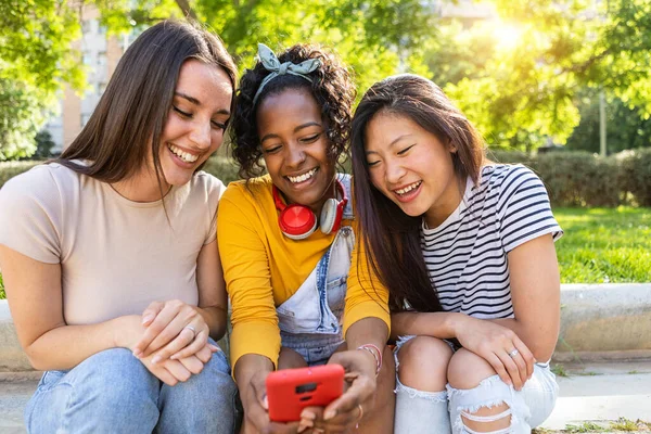 屋外に座って携帯電話を使用して3人の若い多人種の女性の友人 スマートフォンデバイス上でソーシャルメディアコンテンツを共有100万人と女性の友情の概念 — ストック写真