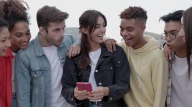 Dışarıda cep telefonuyla gülen genç bir grup mutlu insan sosyal medya uygulamasında komik videolar izlerken gülümseyen çok ırklı öğrenci arkadaşlar. Teknoloji ve gençlik kavramı