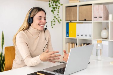 Genç yetişkin bir kadın, müşteri hizmetlerinde dizüstü bilgisayarı kullanarak müşteriyle konuşuyor. Çağrı merkezinde telefon kulaklığıyla çalışan gülümseyen kadın operatör