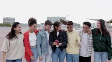 Dışarıda cep telefonu tutan mutlu gülümseyen bir grup genç arkadaş. Ergen çok ırklı insanlar, akıllı telefon ağı uygulamasında komik sosyal medya içeriklerini izlerken birlikte eğleniyorlar.. 