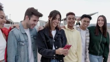 Genç çok ırklı arkadaşlar dışarıda cep telefonu ekranlarına bakarak eğleniyorlar. Çok ırklı gençler birlikte gülüp akıllı telefon ağı uygulamasından komik sosyal medya içeriklerini izliyorlar.. 