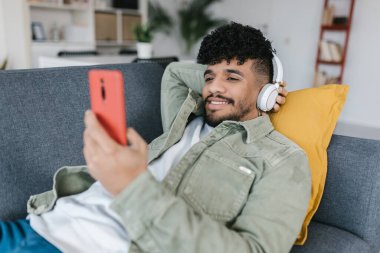 Latin Amerikalı genç adam kanepede oturup cep telefonundan sosyal medyayı izliyor. Teknoloji ve milenyum insanları kavramı