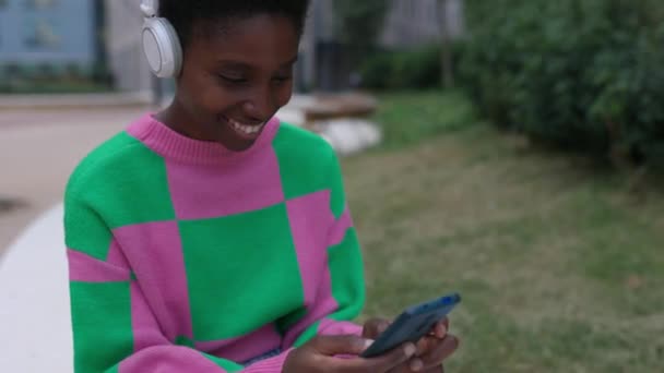 屋外に座っているスマートフォンを使って 若いトレンディーな大人のアフリカ人女性のビデオ テクノロジーとソーシャルメディアの概念 — ストック動画