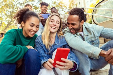 Bir grup uluslararası arkadaş cep telefonu uygulamasından sosyal medya içeriğini izlerken birlikte gülüyorlar. Çeşitli genç yetişkin insanlar dikey selfie portresi çekerken birlikte gülüyorlar.