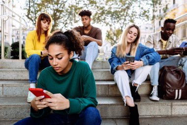 Açık havada akıllı telefon kullanan çok ırklı genç arkadaşlar. Milenyum çeşitliliğindeki yetişkin insanlar cep telefonlarına bağımlılar ve sosyal medya içeriğini izliyorlar.