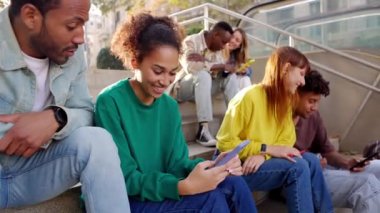 Açık havada oturup akıllı telefondan komik videolar seyreden farklı öğrenci arkadaşlar birlikte gülerken mutlu olun. Genç çok ırklı insanlar sosyal medya cep telefonu uygulaması kullanarak eğleniyor. 