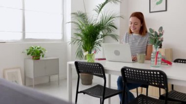 Evde dizüstü bilgisayarda çalışan gülümseyen genç bir yetişkin kadın. Oturma odasındaki bilgisayarla çalışan beyaz bir kadın. Uzak çalışma ve eğitim kavramı.