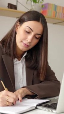 Dizüstü bilgisayarda çalışan genç ve güzel bir iş kadınının iş yerinde not defterine yazdığı dikey çekim videosu.. 