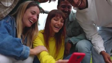 Genç çok ırklı arkadaşlar dışarıda cep telefonu ekranlarına bakarak eğleniyorlar. Çok ırklı gençler birlikte gülüp akıllı telefondan komik sosyal medya içeriklerini izliyorlar. Kentsel yaşam tarzı konsepti