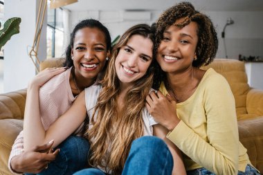 İç mekandaki kameraya gülümseyen çok ırklı kadın arkadaşlarının portresi. Mutlu melez genç kadınlar kanepede oturup birbirlerini kucaklıyorlar.