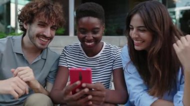 Happy young people having fun sharing social media content on mobile phone. Three multiracial best student friends laughing while using smartphone device app outdoors. Technology lifestyle concept